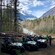 A man removes a helmet while standing near 3 ATVs in a muddy clearing near snow covered mountains